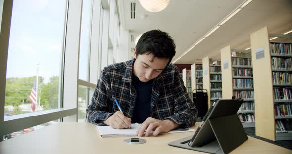 College Student Sitting in Library, Writes in a Notebook, Then Looks at the Camera and Smiles