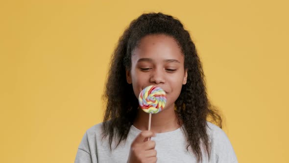 Studio Portrait of Positive African Teen Girl Licking Rainbow Lollipop Enjoying Sweets and Smiling