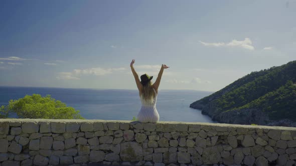 Young woman sitting on a wall with raised arms and enjoying view