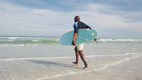 African american senior man walking on a beach holding surfboard and running into the sea