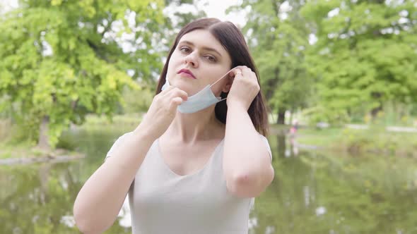 A Young Caucasian Woman Puts on a Face Mask and Looks at the Camera in a Park