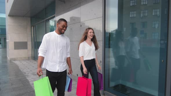 Young Positive Multiethnic Couple Leaving Store Together with Purchases Walking in City Outdoors