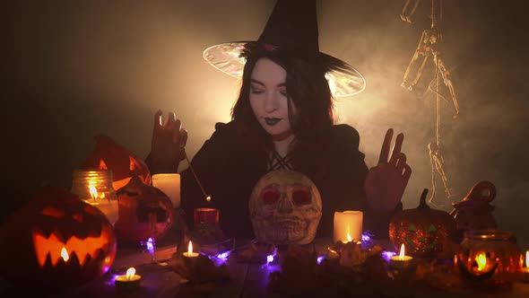 Young Witch in Hat and Dark Mantle is Doing Magic Over Skull Among Candles and Pumpkins