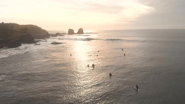 Aerial drone shot at the sea at golden hour flying above surfers with giant rocks on the background,