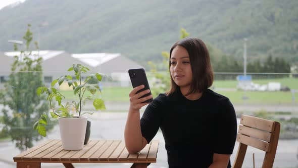 Lonely Thai woman looking at her phone in her urban garden in loud city traffic