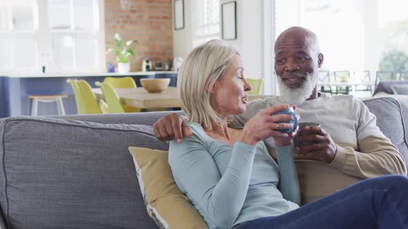 Mixed race senior couple drinking coffee together while sitting on the couch at home