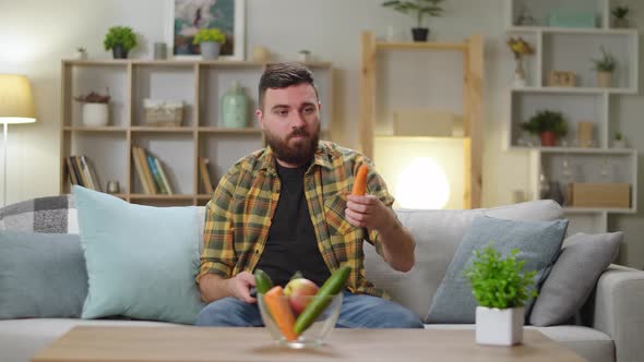 Young Handsome Man Sitting on the Couch at Home and Watching TV and Eating Carrots