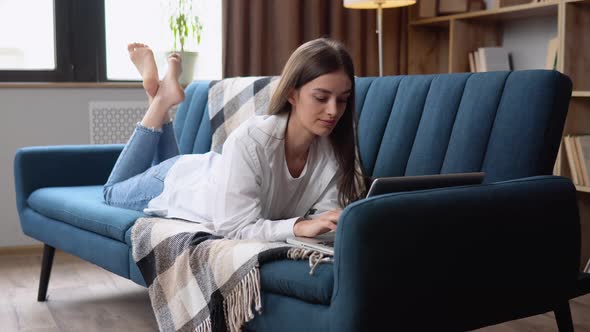 Young Woman Lying on Her Stomach a Sofa Using a Laptop Computer to Surf the Internet with a Happy