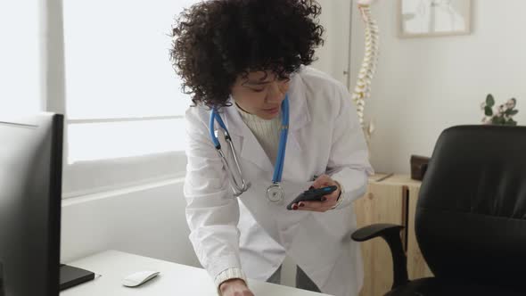 Young Latina Doctor Using Cellphone While Working on Laptop in the Consultation