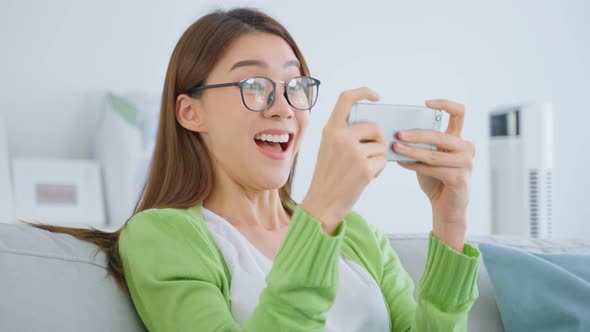 Asian woman wear eyeglasses play mobile game on smartphone at home.