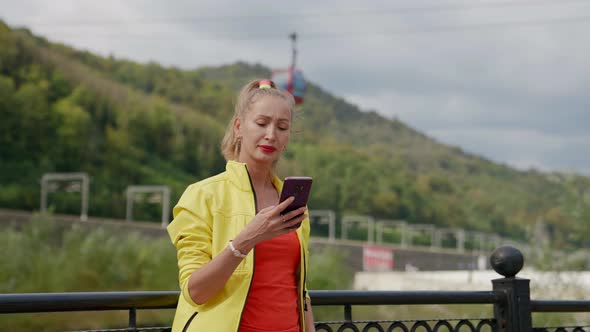 a Woman in Red and Yellow Clothes Stands with a Phone in Hands Against the Background of Blurred