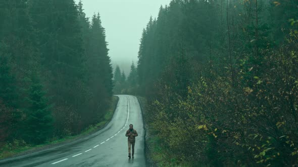 Man Hitchhiker Traveler with Backpack Walks Alog Road Outdoors in Cold Wet Foggy Autumn Day and