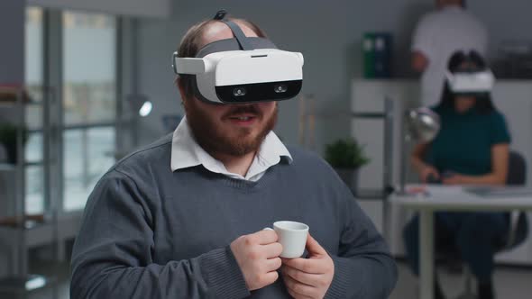 Overweight Businessman in Virtual Reality Goggles Have Video Conference and Drink Coffee