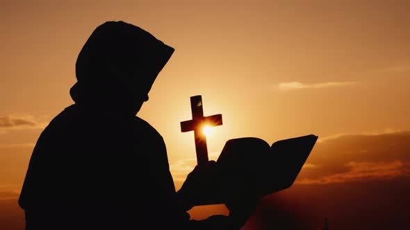 A Monk in a Hood with a Crucifix in His Hands Stands Against the Backdrop of a Dramatic Sky alt