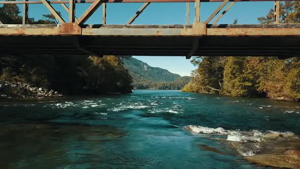 Drone passes under a metal bridge very close to the water of a fast-flowing river.