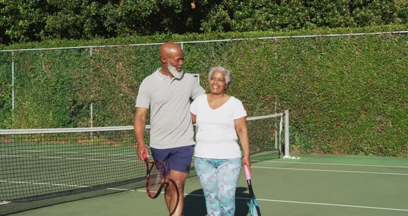 African american senior couple holding rackets walking on the tennis court