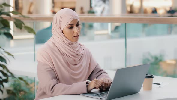 Serious Arab Woman in Hijab Working Remotely Typing on Laptop Writing Book Article Muslim Writer