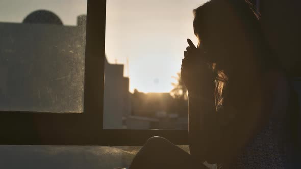 Woman Sits at Window Drinks Coffee Against Sunrise