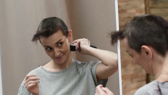 Woman stands in front of a mirror and shave hair bald with the back of head with an electric machine