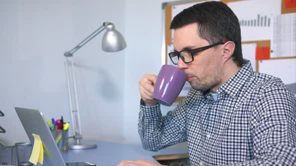 Male Computer User Typing on Keyboard, Looking at the Screen and Drinking Coffee