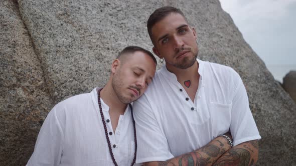 Gay Couple with Closed Eyes Resting Leaning on Each Other Shoulder By the Rock Looking at the Camera