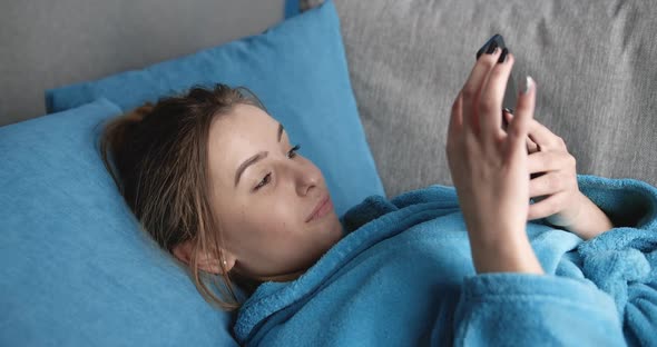 Smiling Girl in Bathrobe Texting with Smart Phone on Sofa