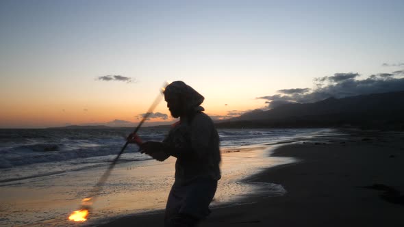 A male fire dancer or performer in silhouette spinning a flaming staff on the beach at sunset with o