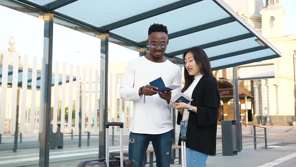 Man Standing on Bus Stop Together with His Cute Satisfied Asian Female Friend and Looking at Tickets