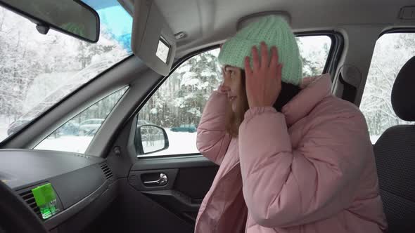 Girl in the Car in Winter Hat and Winter Down Jacket Looks in the Top Mirror Straightens Her Hair
