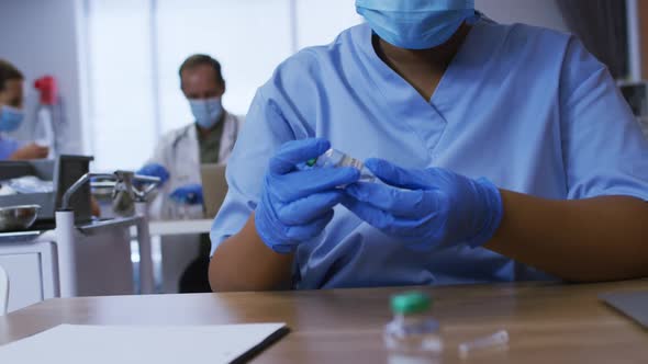 African american female doctor wearing face mask inspecting vial of covid vaccine in hospital