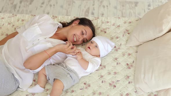 Young Happy Mom Is Playing with a Newborn Baby in the Bedroom on the Bed.