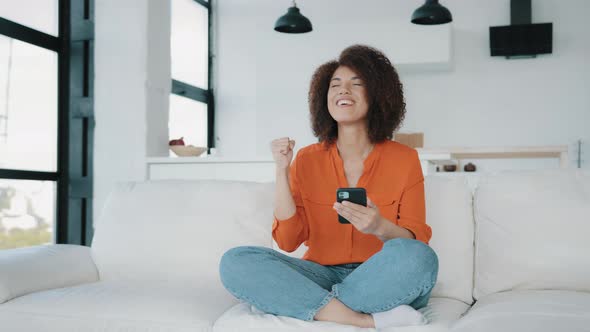 African American Woman Sitting Home Couch Looking Smartphone Using Phone Play Mobile Game Win
