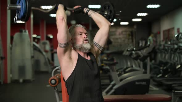 Gray-haired adult man doing french press with a barbell while sitting on a bench in the gym