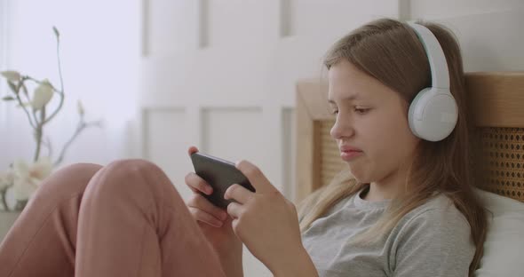 School Girl Is Chatting Online By Smartphone and Listening Through Headphones, Staying Home in Her