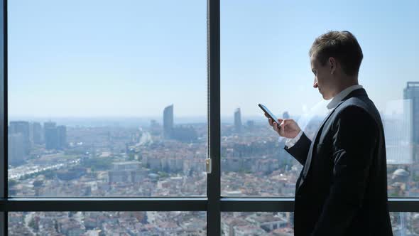 Young Businessman in Office Is Browsing Phone Near the Window with Panoramic City View.