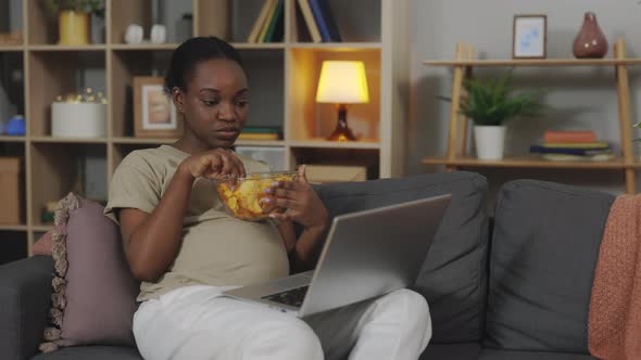 Pregnant Woman Eating Chips and Using Laptop at Home