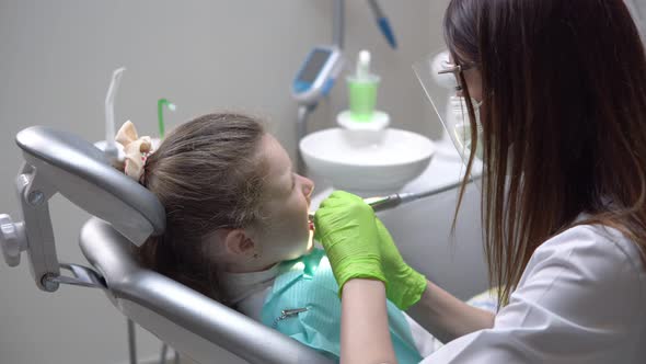 Female Dentist Drilling Tooth of Cute Little Girl at the Dentist Cabinet and Uses Saliva Ejector