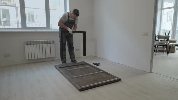 A Builder in a Work Overalls is Assembling a Table in a New Apartment