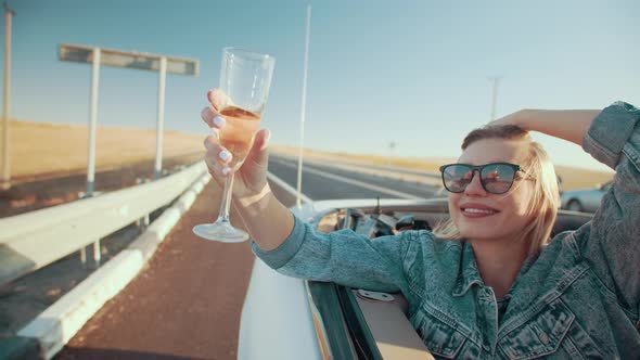 Happy Woman in Denim Jacket and Sunglasses Riding in Convertible Leaning Out Window Holding Glass of