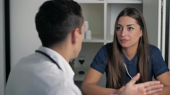 Serious Focused Female Physician in Medical Darkblue Coat Sitting at Table Consulting Male Doctor