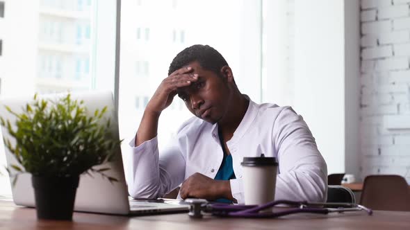 Tired African Black Male Doctor Working on Laptop, Thinking About Problem and Drinking Coffee.
