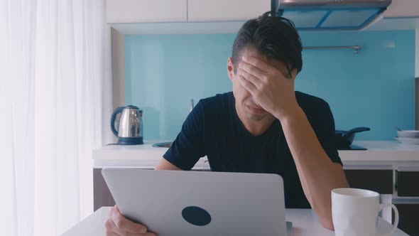 Depressive Young Man Working on a Laptop at Home in the Kitchen