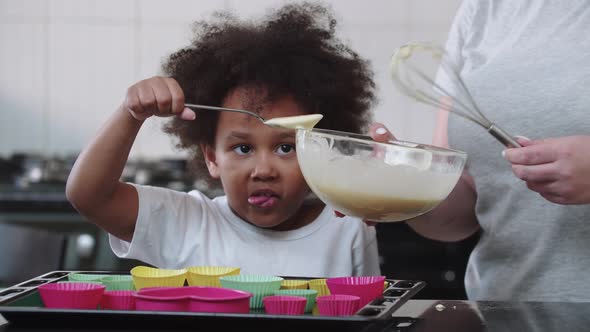 A Little Funny Black Girl Pouring Raw Liquid Dough in the Cake Moulds with a Spoon