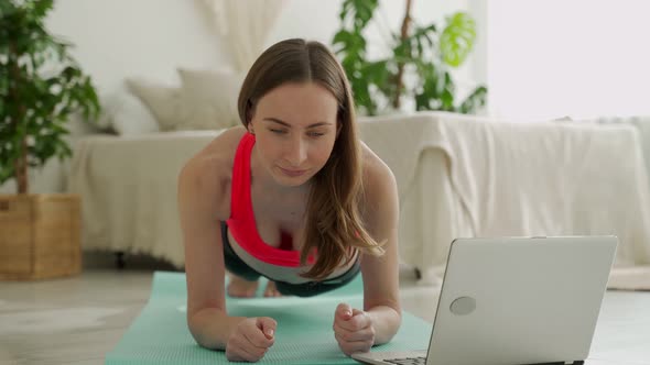 Woman in Sportswear Makes Plank at Mat on Floor and Looks at Laptop of Living Room