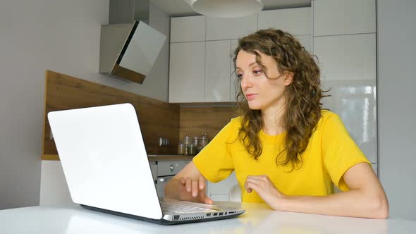 Young Woman with Curly Hair and Yellow Shirt is Working From Home Using Her Laptop at the Kitchen