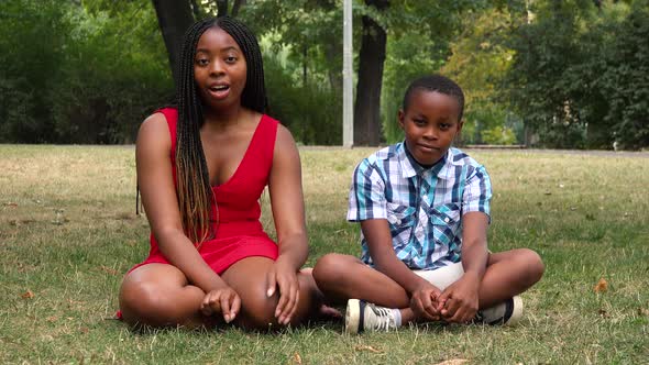 A Young Black Mother and Her Son Sit on Grass in a Park and Talk To the Camera