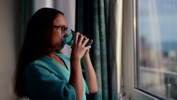 A Pretty Adult Woman with Glasses Standing at the Window of the House in the Morning in a Bathrobe