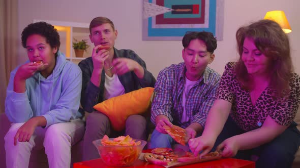 Multiethnic Young Friends Eating Pizza and Chips Watching Tv in Modern Living Room