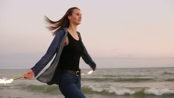 Young Beautiful Woman Running By the Sea During Sunset and Holding Burning Sparkling Candles in Both