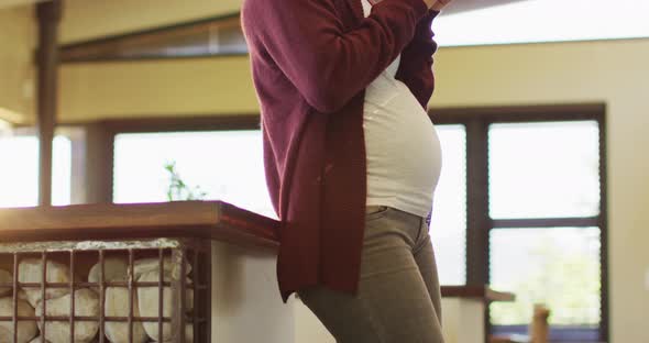 Midsection of caucasian pregnant woman standing in kitchen, touching belly and drinking tea